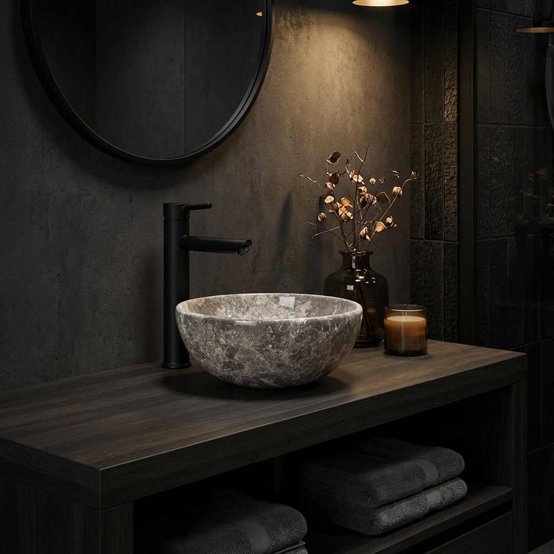 Ronde natuurstenen opzetwastafel met grijsbruine nerf op een donkerhouten console in de sfeervol verlichte badkamer, gecombineerd met zwart beslag, spiegel en subtiele accessoires