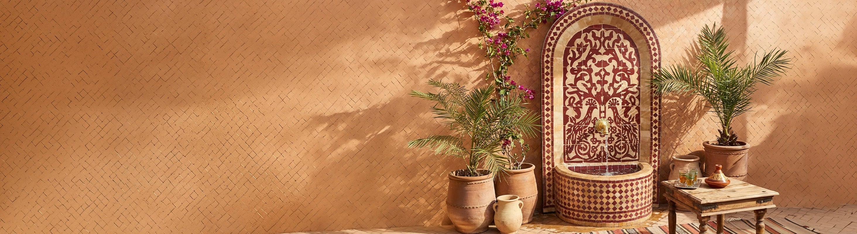 Mosaikbrunnen – Marokkanischer Mosaikbrunnen mit Zellige-Fliesen als Wasserbrunnen im Garten vor warmer Wand mit Pflanzen und orientalischer Atmosphäre