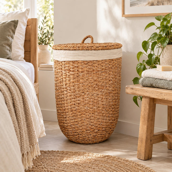 Wasmand met deksel van natuurkleurig zeegras en een lichtgekleurde stoffen inzet, geplaatst tussen het bed en de houten bank in de slaapkamer.