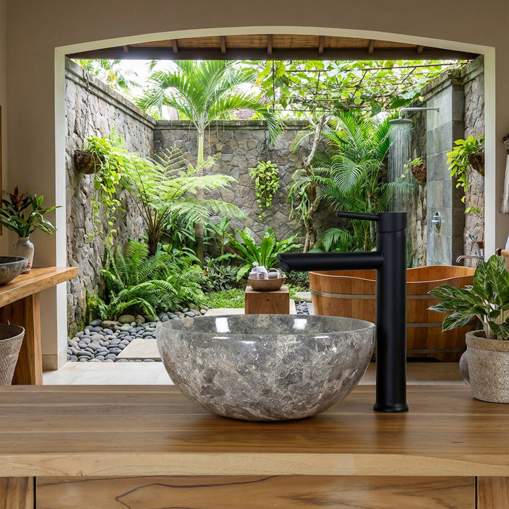 Ronde natuursteen Wastafel met grijsbruine nerf op een houten wastafel in de open spa badkamer met uitzicht op een groene binnentuin, gecombineerd met een zwart vrijstaand armatuur, natuurstenen muur en houten ligbad